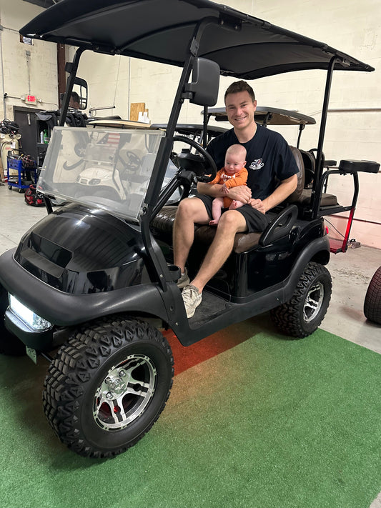 Family-owned golf cart business in Dunedin, Florida – parent holding baby in shop beside a custom Club Car golf cart at American Retrofits.